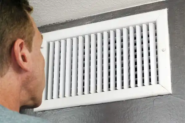 A person examines a white air vent on a gray wall, contemplating the benefits of air duct sealing for improved efficiency.