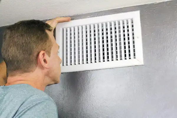 A person carefully inspecting a white wall vent with horizontal and vertical slats, ensuring precise air duct installation.