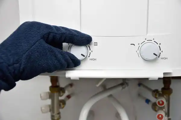 A gloved hand expertly adjusts the temperature knob on a tankless water heater, with visible pipes and control dials confidently set to 'High'—a key step in routine maintenance.